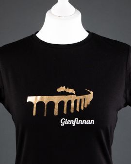 The tshirt on a dressmaker's model. It is black with a high neck and short sleeves. On the front is the Glenfinnan Viaduct with a  steam train passing over, it in gold. Underneath is written 'Glenfinnan.'