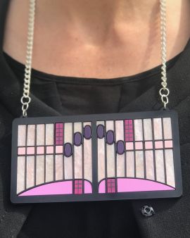 On a woman's neck hangs a silver chain with a large, square, pink pendant. It has a black border and features a design of lines and circles, inspired by a light at the Hill House. 